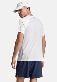 Camiseta atlética blanca de manga corta y tela texturizada, combinada con pantalones cortos azul marino que tienen aberturas laterales. Los accesorios incluyen una gorra blanca.