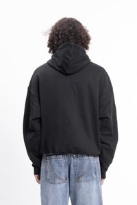 Personne avec des cheveux bouclés portant un sweat à capuche noir et un jean en denim bleu clair, se tenant de dos contre un fond blanc uni.