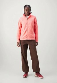 Žena u koraljnoj jakni na zatvaranje, smešnim hlačama i crvenim patikama stoji nasuprot jednostavne svetle pozadine.