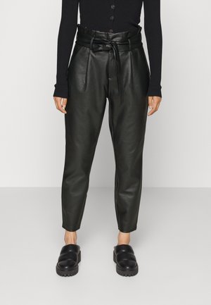 Kobieta ubrana w czarne, wysokiej talii spodnie z ekologicznej skóry, z paskiem do wiązania, w połączeniu z czarnymi butami wsuwanymi i czarną, długorękawową bluzką.