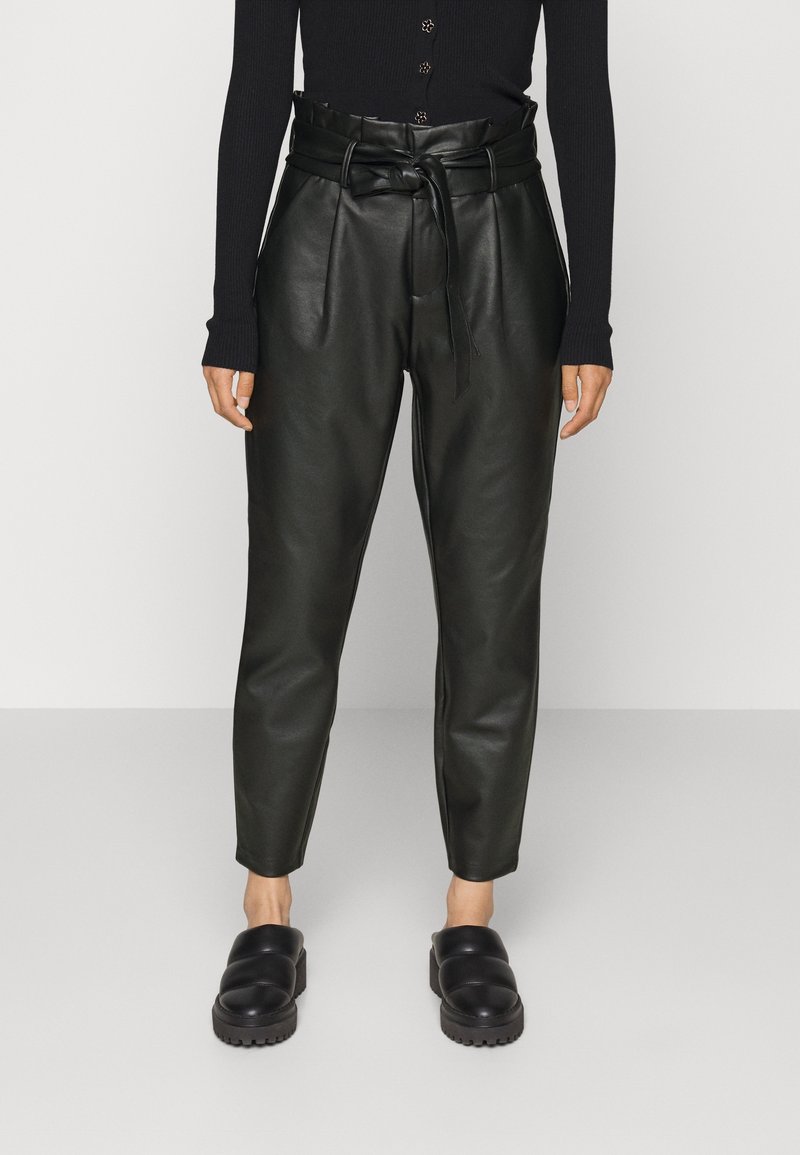 Femme portant un pantalon noir en similicuir taille haute avec une ceinture nouée, associé à des chaussures noires plates et un haut noir à manches longues.