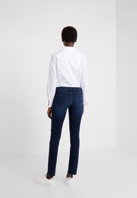 Camisa blanca de manga larga combinada con jeans de color azul oscuro, con un corte ajustado, tiro medio y dos bolsillos traseros. Zapatillas blancas completan el conjunto.