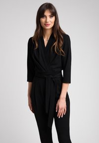 Czarny kombinezon z rękawami o długości trzy czwarte, wiązanym w pasie i miękkiej fakturze. Prosty design bez wzorów i widocznych elementów metalowych.