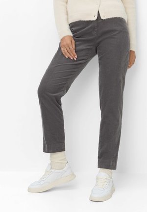 Personne portant un pantalon droit gris, des chaussettes crème, des baskets blanches et un pull crème, la main reposant sur la cuisse.