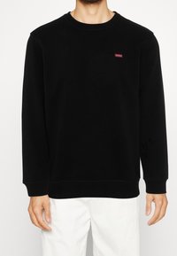 Sudadera negra de cuello redondo hecha de una mezcla de algodón. Presenta un pequeño logo rojo de "GUESS" en el pecho y puños y dobladillo acanalados.
