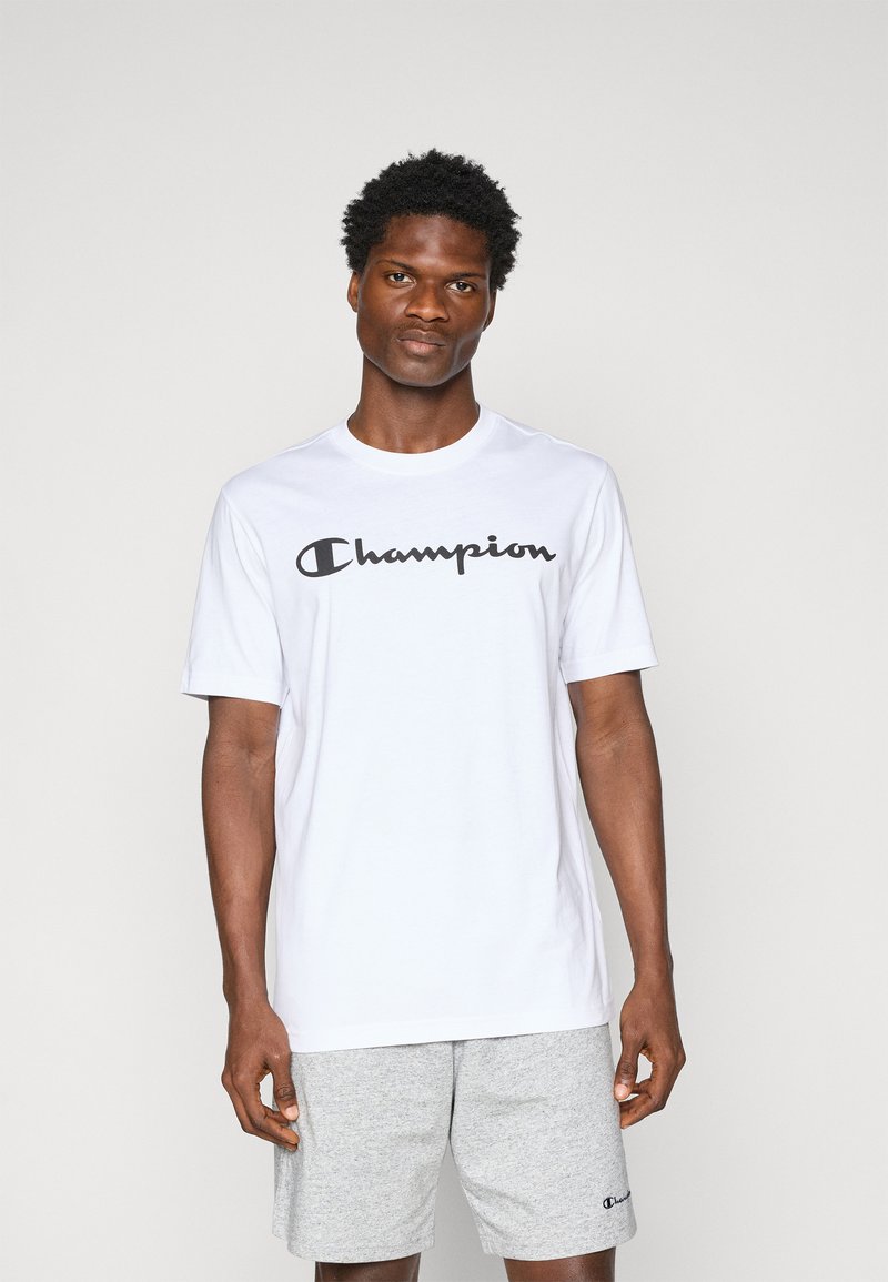 Weißes Kurzarm-T-Shirt aus Baumwolle mit einem schwarzen "Champion"-Logo über der Brust; kombiniert mit grauen Shorts.