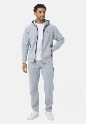 Hombre de pie vestido con una sudadera con capucha gris clara con cremallera, pantalones deportivos a juego, camiseta blanca y zapatillas blancas sobre un fondo blanco liso.