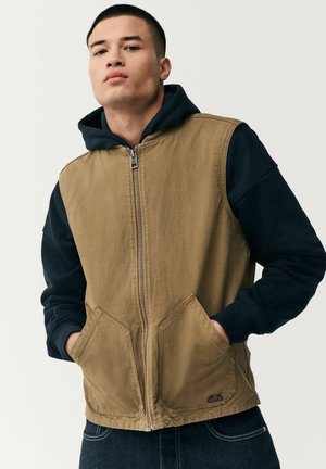 Brown, zip-up vest with pockets over a black hooded sweatshirt. Features a sturdy fabric and contrasting textures in the layered design.