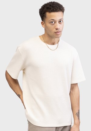 Chemise courte à manches courtes de couleur crème, texturée, avec un col rond et une coupe décontractée, présentant un motif gaufré subtil et des bords lisses.