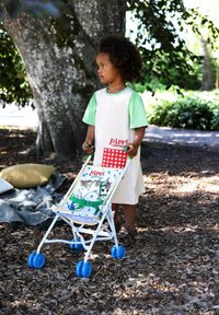 Legetøjsklapvogn med en hvid ramme og blå hjul, dekoreret med et mønstret stof med et tegnefilmsdesign. Barnet i en lysbeklædning holder den.