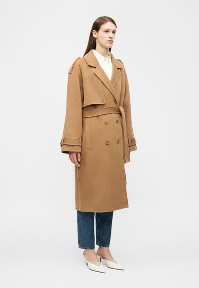 Donna che indossa un cappotto lungo in lana color cammello con cintura, sopra jeans blu e scarpe bianche con tacco, in piedi davanti a uno sfondo chiaro e semplice.