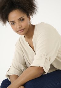 Beige Leinenbluse mit V-Ausschnitt und hochgerollten Ärmeln, kombiniert mit dunkler Jeans. Klare Haut und dunkel lockiges Haar, lässig gestylt. Natürliches Licht.