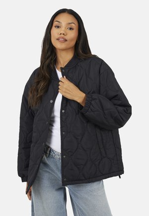 Kobieta ubrana w luźną czarną pikowaną kurtkę na białej koszuli i jasnoniebieskich dżinsach, trzymająca kurtkę przy kołnierzu na jednolitym tle.