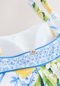 Vestito bianco con motivo floreale blu e giallo, con spalline sottili e una piccola fibbia decorativa dorata sulla scollatura.