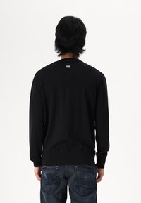 Schwarzes Longsleeve-Shirt mit rundem Ausschnitt und gerippten Bündchen. Auf der Rückseite befindet sich ein kleines Logo-Tag oberhalb des Ausschnitts. Kombiniert mit dunklen Denim-Jeans.