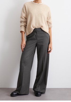Femme portant un pull beige aux manches retroussées, un pantalon large à rayures fines gris foncé et des chaussures noires, debout contre un mur blanc.