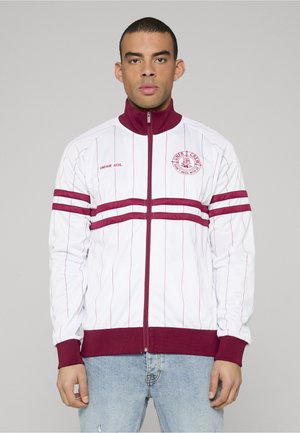 Muž v bielej vetrovke so zipsom, ktorá má tenké červené vertikálne pruhy, červené horizontálne pásy na rukávoch a na hrudi, a modré rifle, pozerajúci sa vpred.