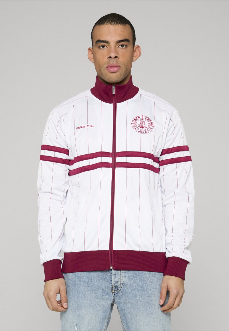 Hombre con chaqueta blanca con cremallera con finas rayas verticales rojas, bandas horizontales rojas en las mangas y el pecho, y jeans azules, mirando hacia adelante.