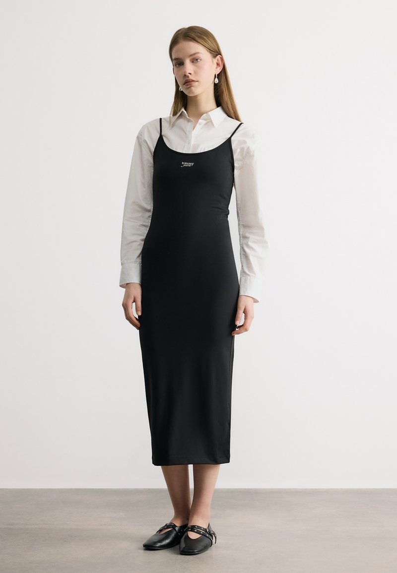 Γυναίκα που φοράει μαύρο midi φόρεμα χωρίς μανίκια πάνω από λευκό πουκάμισο με γιακά, συνδυασμένο με μαύρα παπούτσια Mary Jane, όρθια μπροστά σε απλό φόντο.