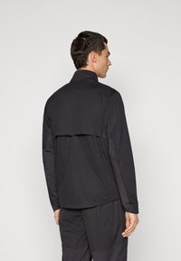 Man facing away wearing a black long-sleeve jacket with a back flap and matching black pants against a plain background.