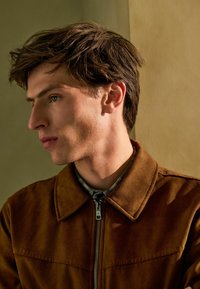 Chaqueta de ante marrón con cremallera y cuello ancho, llevada sobre una camiseta gris texturizada; vista de perfil contra un fondo neutro y suave.