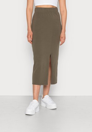 Kvinde iført olivengrøn midi blyantsnørk med frontslids, hvid cropped top og hvide platåsneakers på ensfarvet baggrund.