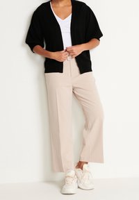 Osoba ubrana w beżowe szerokie spodnie z paskiem, białe trampki, białą bluzkę oraz czarny kardigan z odkrytym przodem stoi przy białej ścianie.