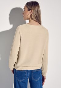 Felpa beige con un fit rilassato, texture a costine e maniche lunghe, abbinata a jeans blu con cuciture accentuate e tasche posteriori.