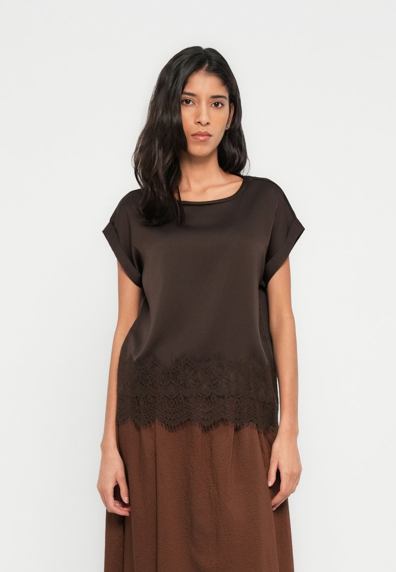 Καφέ κοντομάνικη μπλούζα με λεία υφή, που διαθέτει λεπτομέρεια δαντέλας στο τελείωμα. Η μπλούζα έχει στρογγυλή λαιμόκοψη και πέφτει χαλαρά πάνω από μια καφέ φούστα.