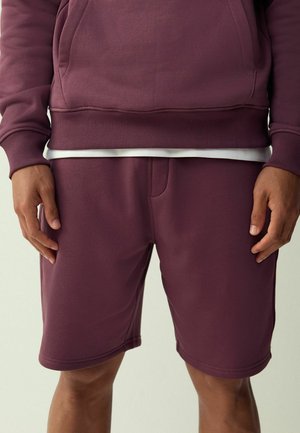 Mężczyzna w bordowych szortach i pasującej bluzie z białą koszulą wystającą na wysokości talii, stoi z rozluźnionymi rękami.