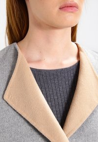 Närbild av en person som bär en grå ribbad topp och en grå kappa med en ljusbeige bred krage, där deras hals och läppar syns.