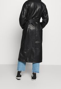 Manteau long en cuir noir avec une taille ceinturée, présentant une texture lisse et un détail de couture minimaliste. Porté par-dessus un jean bleu à jambes larges et des baskets noires.