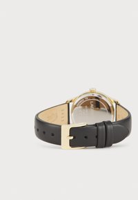 Correa de reloj de piel negra con hebilla dorada, presenta una caja redonda y un fondo de cristal claro que muestra la marca y los detalles del modelo.