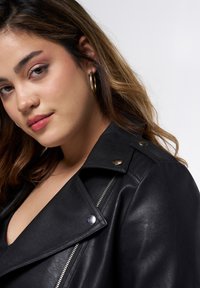 Veste de moto en cuir noir avec quincaillerie argentée, fermeture éclair et épaulettes. Le modèle porte un maquillage léger et des boucles d'oreilles créoles.
