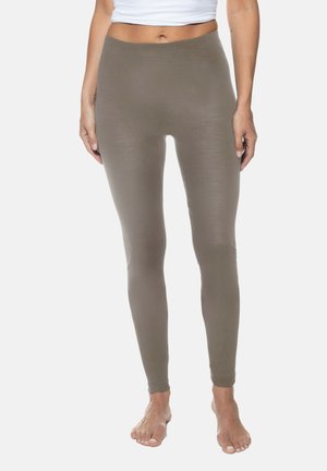 Frau, die taupefarbene, gerippte Leggings und ein weißes, bauchfreies Oberteil trägt, barfuß vor einem schlichten, hellen Hintergrund steht.