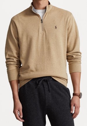 Mies, jolla on beige puolivetoketjullinen neule valkoisen paidan päällä, tummanharmaat naruvöiset housut ja vasemmassa ranteessa ruskealla nahkahihnalla varustettu rannekello.