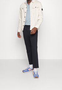 Leren crèmekleurige jack met knopen over een blauwe polo, gecombineerd met zwarte tapered broek en blauwe New Balance sneakers met oranje accenten.