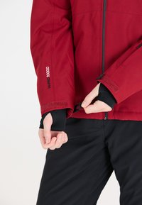 Röd vattentät jacka med svarta detaljer; har justerbara ärmslut med kardborre och tumhål; sömdetaljer och dragkedjestängning.