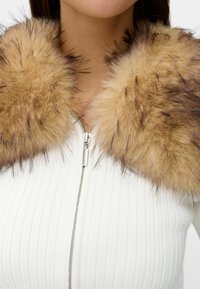 Faux pälskrage i ljusbrun med mörka detaljer, fäst vid ett vitt ribbat zip-up-plagg. Mjuk textur med voluminöst mönster.