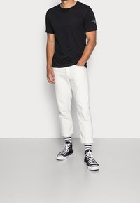 Černé tričko s krátkým rukávem, bílé džíny s ohrnutými nohavicemi, černé vysoké tenisky a černobílé pruhované ponožky. Minimalistický design, volný střih.