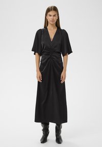 Must jõhvikas satin kleit, millel on sügav V-kaelus, kokkuvooditud esikülg ja voolavad varrukad. Pikad, saledad siluett on rõhutatud kõrgete mustade saapastega.