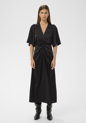 Must jõhvikas satin kleit, millel on sügav V-kaelus, kokkuvooditud esikülg ja voolavad varrukad. Pikad, saledad siluett on rõhutatud kõrgete mustade saapastega.