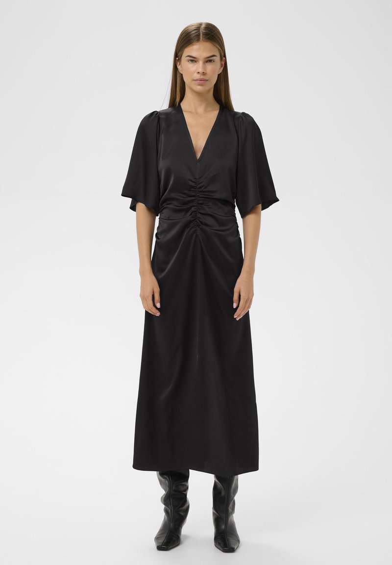 Must jõhvikas satin kleit, millel on sügav V-kaelus, kokkuvooditud esikülg ja voolavad varrukad. Pikad, saledad siluett on rõhutatud kõrgete mustade saapastega.