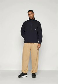 Mand står imod en ensfarvet baggrund iført en sort fleece jakke med 1/4 lynlås, beige løse bukser og sorte sneakers, med den ene hånd i lommen.