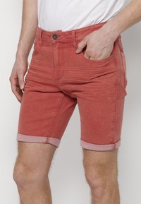 Pantalones cortos de mezclilla roja con puños doblados, diseño de cinco bolsillos, cierre frontal con botón y sutiles detalles de textura. Estilizados con una camisa blanca.