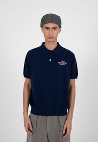 Jeune homme portant une casquette plate grise, un polo bleu marine avec le logo "Prohibited Racing Club" et un pantalon gris plissé, sur un fond clair uni.