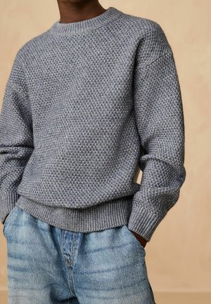 Personne portant un pull en maille gris texturé et un jean en denim bleu clair avec les mains dans les poches, sur un fond beige uni.