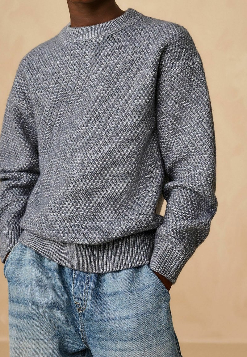 Personne portant un pull en maille gris texturé et un jean en denim bleu clair avec les mains dans les poches, sur un fond beige uni.