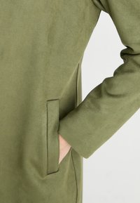 Chaqueta verde oliva hecha de un material suave y texturizado, con un corte holgado, que presenta un bolsillo lateral oculto y detalles de costura en los bordes.
