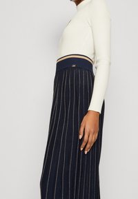 Námořnická plisovaná sukně se zlatými vertikálními pruhy, se žebrovaným pasem a kontrastním béžovým akcentem. Viditelná měkká textilie.